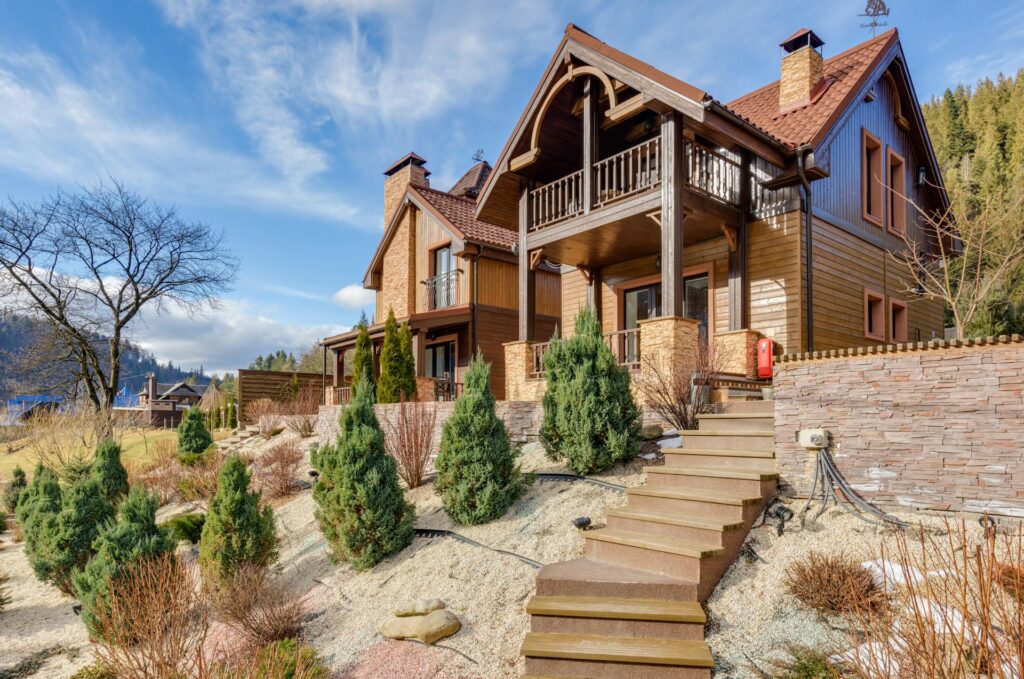 A Large, Two-Story Wooden House With A Red Shingle Roof And Stone Accents, Set On A Landscaped Slope With Small Conifer Shrubs, Rock Mulch, And Brown Wooden Stairs Leading Up To The Front Entrance. The Home Features Balconies, Large Windows, And Is Surrounded By A Mountainous Forest Area Under A Partly Cloudy Blue Sky.