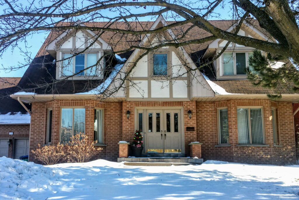 A Large Two-Story Brick House With Tudor-Style Architecture, Snow Covering The Front Yard And Roof, Leafless Tree Branches Extending Across The Top Of The Image, And A Small Decorated Christmas Tree By The Front Door.