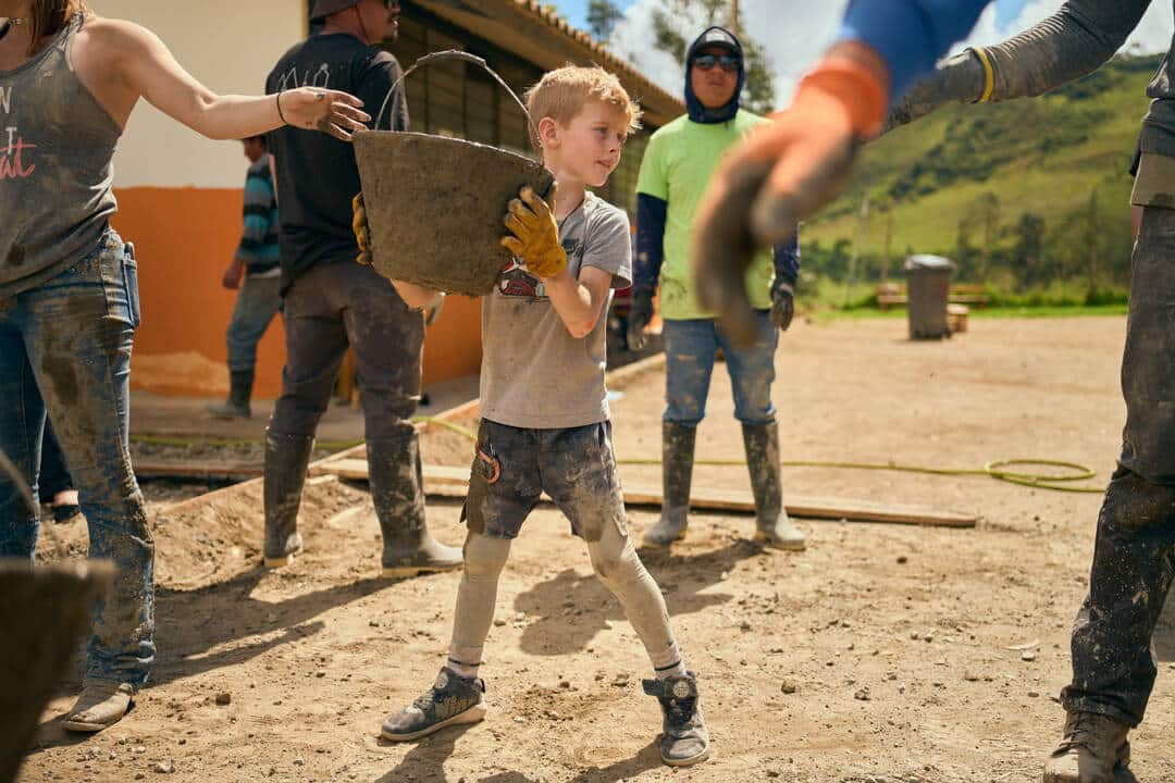 A Young Boy Participates In A Community Service Project, Carrying A Bucket With Other Volunteers In The Background. A Young Boy Participates In A Community Service Project, Carrying A Bucket With Other Volunteers In The Background.